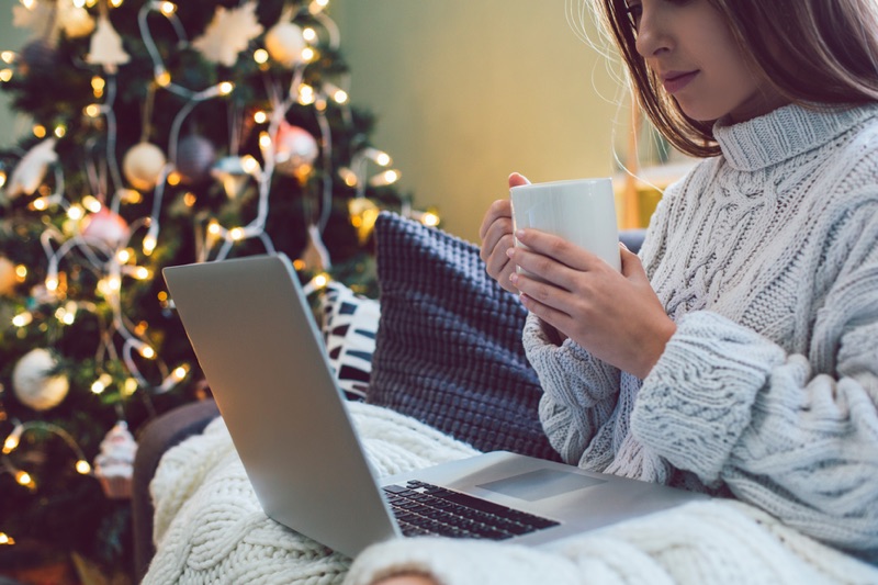 Young woman holding a mug and reading on her laptop and sitting on couch by the Christmas tree.